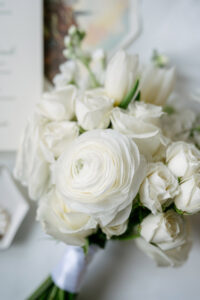 White rose and ranunculus bouquet wrapped in ribbon, with a blurred invitation and tray in the New Orleans wedding scene.