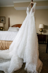A white sleeveless wedding dress with deep V neckline and full skirt hangs from a bedpost in a hotel room.