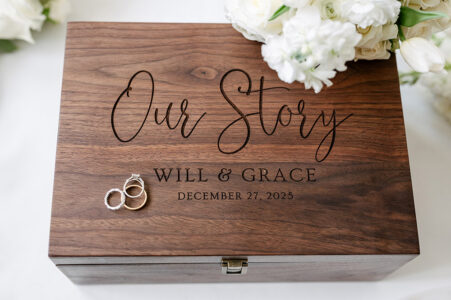 Wooden keepsake box engraved with Our Story, Will & Grace, December 27, 2025; wedding rings and white flowers on top.