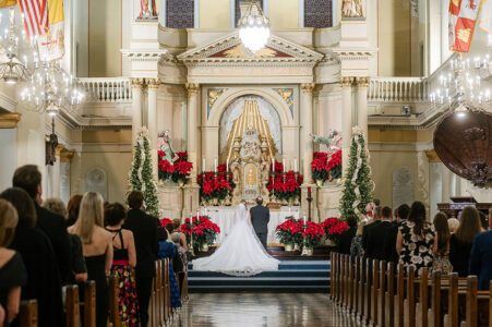 Bride and groom kneeling at altar in decorated church with guests, red poinsettias, white lights, and golden accents.