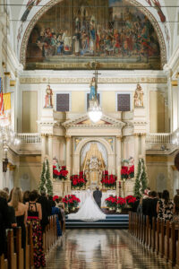 Bride and groom at altar in ornate church with red flowers, guests in pews, under a painted dome—classic Gulf South wedding.