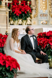 Bride and groom smile amid red poinsettias in a gold-accented Louisiana church, highlighting wedding elegance.