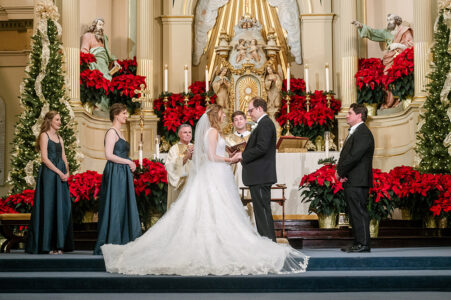 Bride and groom at church altar with bridesmaids, groomsman, and Christmas decor including red poinsettias—festive wedding inspiration.
