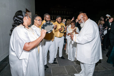People in white and gold play music and clap outdoors at night, creating a joyful Louisiana wedding atmosphere.