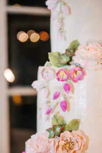 White wedding cake with pink and white sugar flowers, green leaves, and blurred lights in the background.