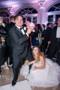A man in a tuxedo shakes a maraca as a smiling bride kneels at a lively Louisiana wedding with well-dressed guests.