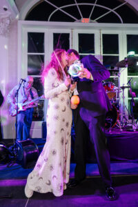 A woman in a sequined dress and a man in a suit smile on stage at a lively New Orleans wedding with a band and colorful lights.