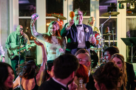 Bride and groom celebrate with maracas onstage at a lively New Orleans wedding, guests dancing with live band in background.