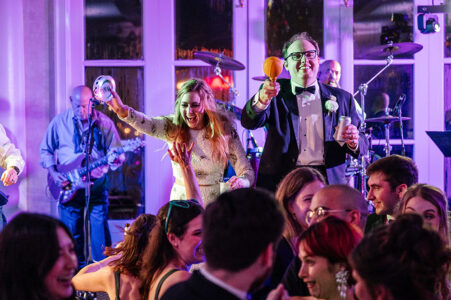 A joyful couple in formal attire play maracas on their wedding dance floor, surrounded by smiling guests and a live band.