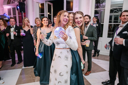 Two women in formal dresses laugh and hug at a lively New Orleans wedding, holding glowing cups as guests celebrate in the background.