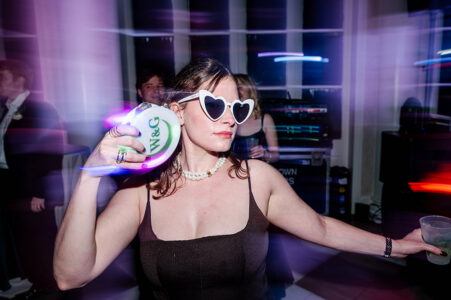Woman in heart-shaped sunglasses holding a decorated drink at a lively New Orleans wedding party with colorful lights.