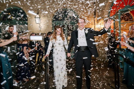 Smiling couple in formal wear walks hand-in-hand through confetti, cheered by guests at a lively Gulf South wedding.