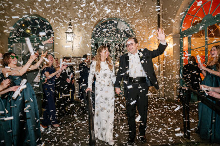 Newlyweds smile, walking hand in hand as guests throw white confetti outside a brightly lit building at night.