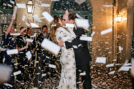 A couple in formal wear kisses under falling confetti, cheered on at a magical Gulf South wedding reception at night.