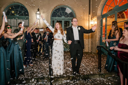 Bride and groom celebrate outside with cheering guests and confetti after wedding; guests smile, take photos on the steps.