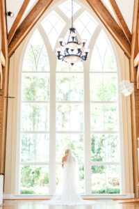 Bride in white dress stands before tall arched windows in an elegant room with wooden beams and a large chandelier.