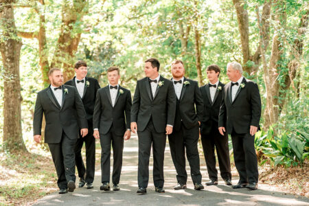 Seven men in black tuxedos and bow ties walk together on a shaded path, smiling and talking among trees—classic wedding inspiration.