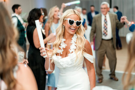 Woman in white dress and heart sunglasses holds champagne and glow stick at lively Louisiana wedding with dancers behind her.