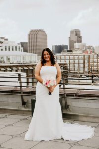 Bride in a white dress smiles on city rooftop, holding pink bouquet after her Louisiana wedding ceremony.