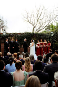 Bride and groom at outdoor Louisiana wedding, with bridesmaids in red, groomsmen in black, and guests seated on white chairs.