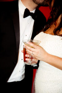 Elegant couple in a white beaded dress and black tuxedo, holding a cocktail against a red background—wedding inspiration.