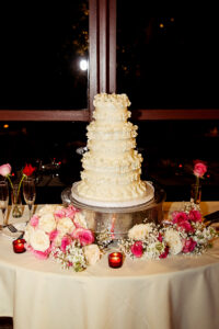 Four-tier white wedding cake with ruffled icing, roses, candles, and champagne flutes on a romantic table setting.