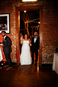 Bride in white dress and groom in tuxedo smile, walking hand-in-hand into a warmly lit wedding reception with brick walls.