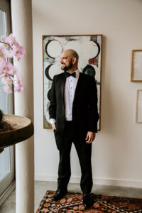 Man in black tuxedo smiles near a window and potted orchid, with art-decorated walls in an elegant New Orleans-style scene.