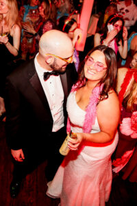 Smiling couple in formal wear and sunglasses at a lively wedding, woman in white dress with pink boa holds a drink under red lights.
