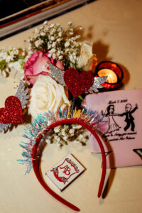Red headband with glittery heart wings, roses, flowers, candle, dancing couple napkin, and AB matchbox on cream surface.
