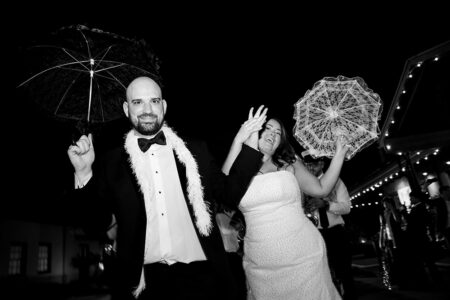 A joyful couple in formal wear wave and hold parasols at a New Orleans wedding, with guests and string lights behind them.