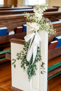 White flowers, greenery, and a white ribbon adorn a church pew; hymnals in the background create classic Gulf South wedding charm.