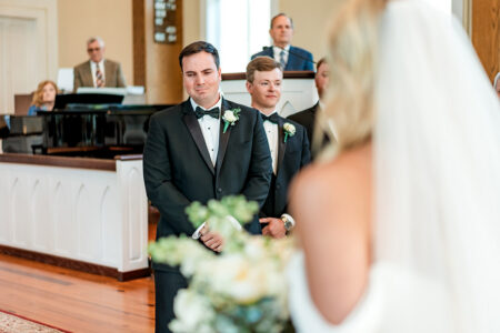 Emotional groom in a tuxedo waits at the altar during a New Orleans wedding as guests look on in a bright church.