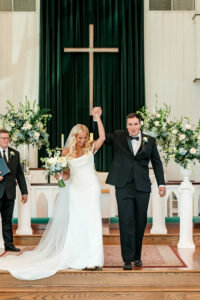 Smiling bride and groom walk down a flower-decorated church aisle, holding hands and raising arms in joyful celebration.