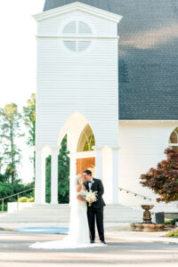 Bride and groom smile outside a white church, dressed for a classic Gulf South wedding, surrounded by greenery and sunshine.