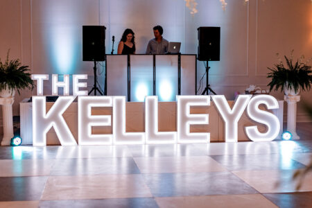 Illuminated letters spell THE KELLEYS before a DJ booth with two people, lush plants, and blue lighting at an indoor reception.