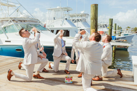 Six men in light suits kneel on a dock by boats, raising bottles as if taking a drink. A beer case sits on the wooden planks between them. It is sunny with a blue sky and scattered clouds.