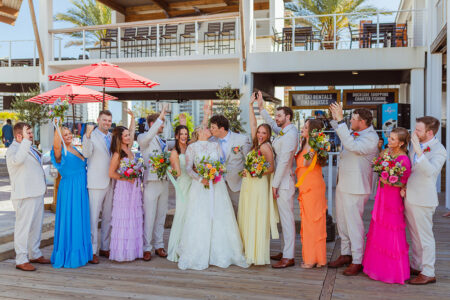 A wedding party cheers as a bride and groom kiss outdoors. Bridesmaids wear colorful dresses, and groomsmen wear light suits. Everyone holds bouquets or raises hands, celebrating under a sunny, open-air venue.