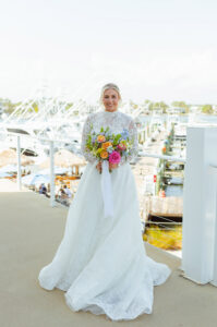 A bride in a white lace gown holds a colorful bouquet, smiling on a balcony overlooking a marina with boats in the background.