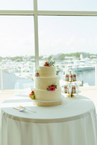 Three-tiered white cake with floral decorations on a round table by a window, overlooking a marina. Nearby are a cake knife, server, three Diet Coke cans, and two glasses.