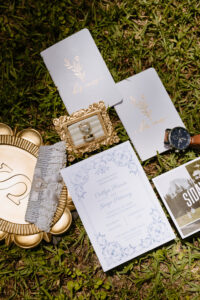 Flat lay of wedding details including invitation suite, rings, heirloom vow books, watch, lace garter, and save-the-date card arranged on grass.