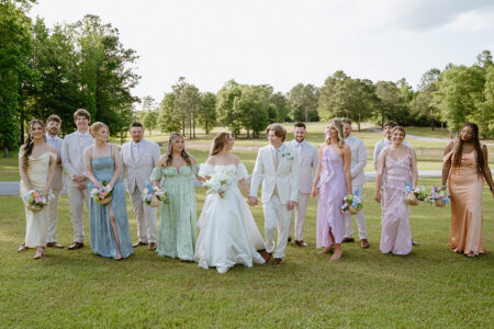 Bride and groom walk across a lawn with their wedding party dressed in soft pastel attire.