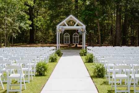 Outdoor wedding ceremony space with white chairs lining a long aisle leading to a flower-adorned white arbor.