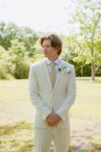 Groom in a light cream suit stands outdoors before the ceremony with hands clasped.